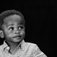 Natural light black and white image of a very young Black boy looking up and to the right on a black background